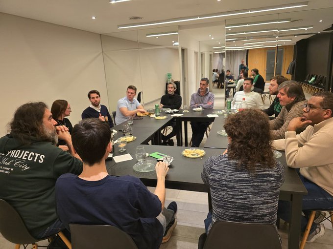 A group of about ten people sit around a long black conference table in a modern office space, with plates and drinks on the table.