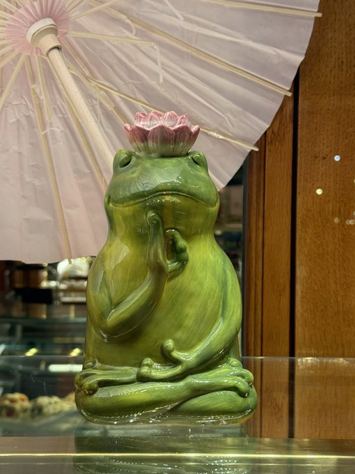 A green ceramic frog figurine sits in a meditative lotus pose with a pink lotus flower on its head, positioned beneath a paper parasol on a glass shelf.