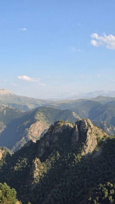 A scenic mountain landscape viewed from a high vantage point shows a rocky peak covered in green forest, with distant mountain ranges stretching across the horizon under a blue sky with scattered clouds.