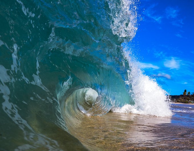 A large, curling ocean wave with blue water and white foam crashing onto a sandy beach.