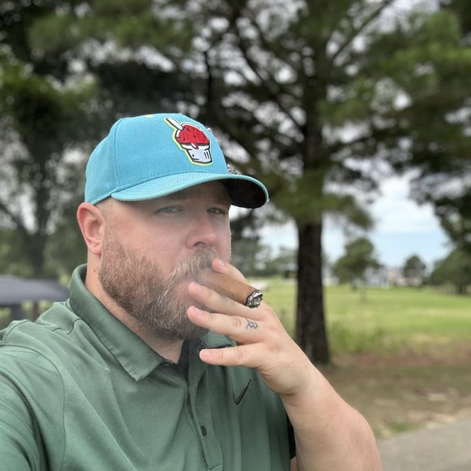 A bearded man wearing a green polo shirt and a teal 'Slurpee' baseball cap smoking a cigar outdoors, with trees and a grassy field in the background.