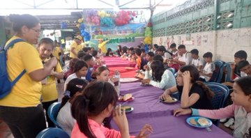 A long, crowded indoor gathering of people, mostly women and children, seated at long tables covered with purple and pink cloths.