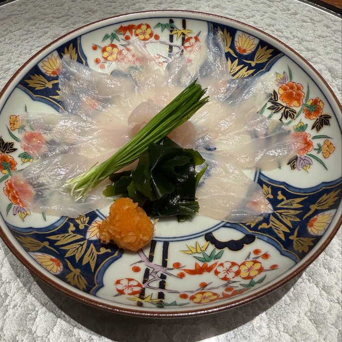 An ornately decorated ceramic plate featuring thinly sliced raw fish sashimi arranged in a fan pattern, garnished with green onions, wakame seaweed, and orange roe.