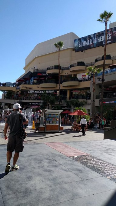 An outdoor shopping plaza scene on a sunny day, with palm trees, a Skechers and Sephora storefront, and a large banner reading 'SUMMER OF HEROES' by Upbound Group.