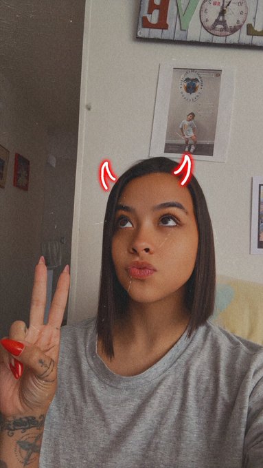 A young woman with a bob haircut makes a peace sign while wearing a gray shirt, with digital devil horns added as a filter over her head.