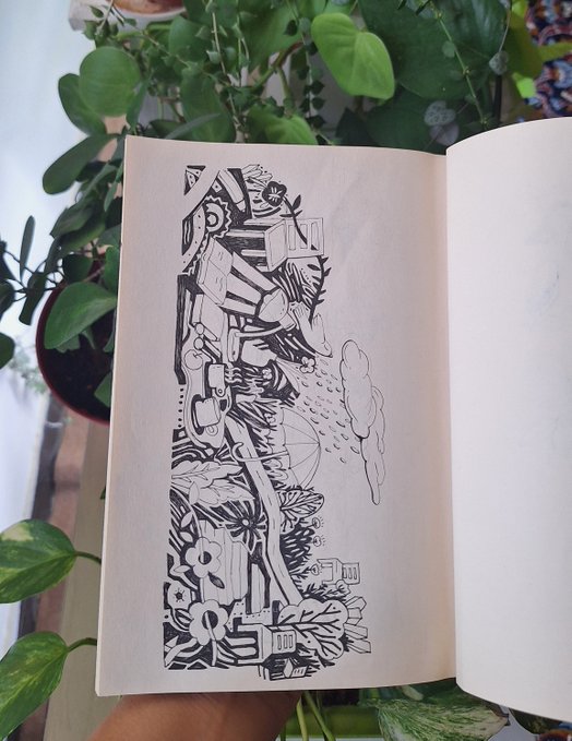 An open sketchbook displays a detailed black-and-white ink drawing of a complex, surreal landscape with structures and flowers, held near green houseplants.