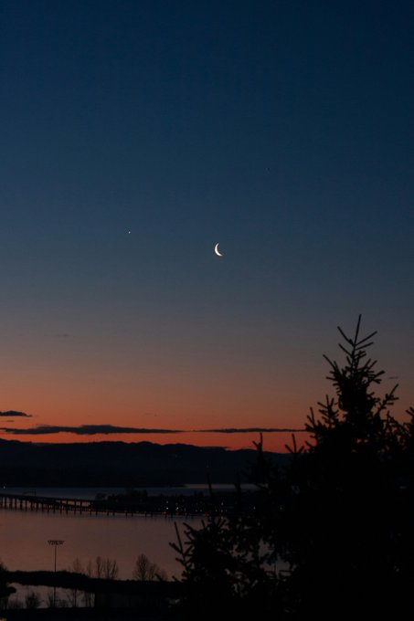 A serene twilight photograph of a crescent moon and a bright celestial body (likely Venus) in a gradient blue-to-orange sky above silhouetted mountains, a body of water, and dark pine trees in the foreground.