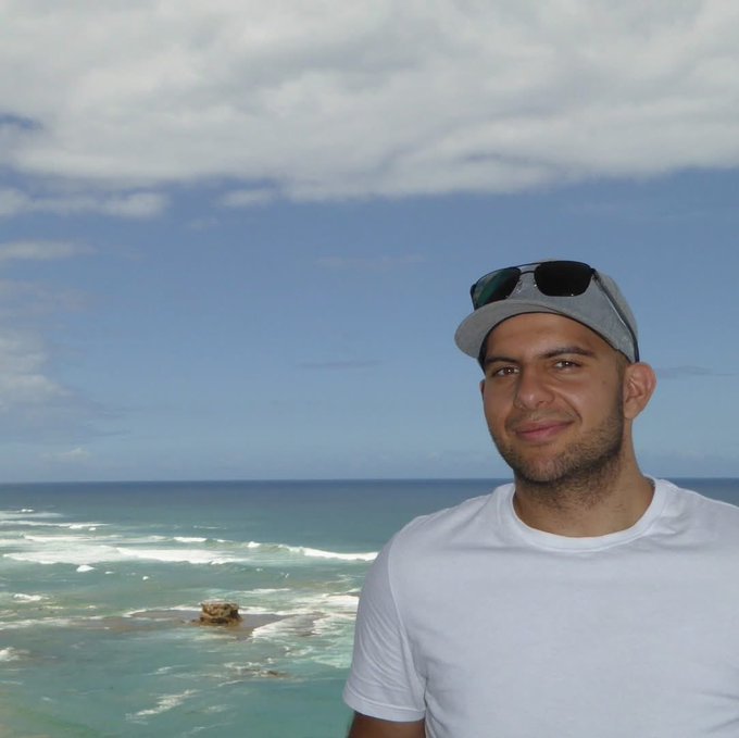 Man in white t-shirt with sunglasses on cap smiling in front of a tropical ocean with turquoise water