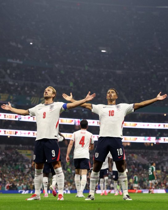In this image, two soccer players from the England national team are seen celebrating on the pitch with their arms outstretched. One player is wearing the jersey with the number 9 and the name "Kane" on the back, while the other player is wearing the jersey with the number 10 and the name "Bellingham" on it. The background shows a stadium filled with cheering fans, and the image appears to be related to the post "Whos talking now @moonpay" by highlighting the performance of these two star players.