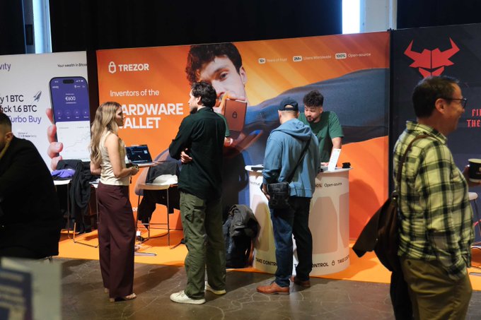 Attendees at a crypto conference booth displaying a large Trezor advertisement reading 'Inventors of the HARDWARE WALLET' with promotional banners in the background.