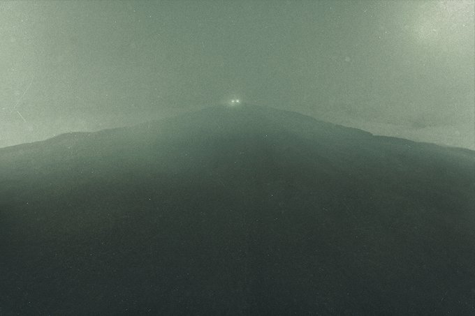 Two glowing lights hover above a dark, misty mountain peak shrouded in a hazy green-grey sky.