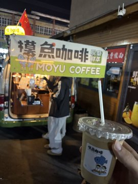 A nighttime street view featuring a brightly lit yellow sign reading '摸鱼咖啡 MOYU COFFEE' above a small cafe counter, with a customer ordering and a hand holding an iced coffee in the foreground.