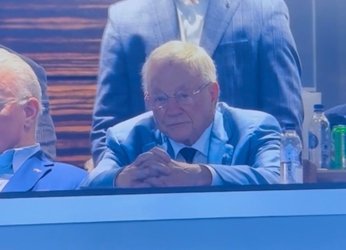 An older man with white hair and glasses, wearing a light blue suit and a striped tie, sitting with his hands clasped at a table, with other people partially visible around him.