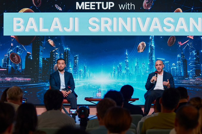 Two men seated on stage in front of an audience at a 'MEETUP with BALAJI SRINIVASAN' event, with a futuristic cityscape and Bitcoin-themed backdrop featuring planets and digital network graphics.