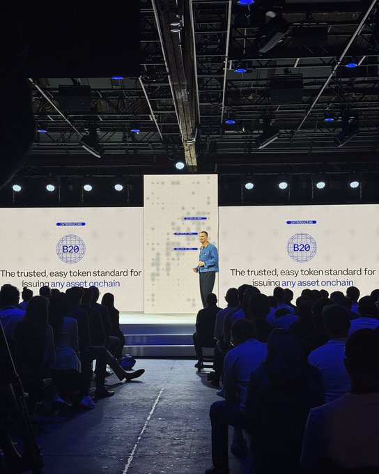 The image shows a man standing at a podium, giving a speech to a group of people who are seated in front of him. There are two large screens behind him, one of which displays a logo that reads "B20 token standard". The audience appears engaged and attentive as they listen to the speaker's presentation on asset tokenization or issuing tokens for an asset chain.