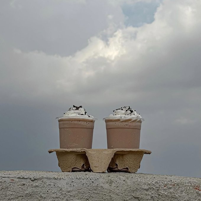 Two clear plastic cups filled with chocolate beverage, topped with whipped cream and dark crumbles, sit in a cardboard holder on a concrete ledge beneath a cloudy sky.