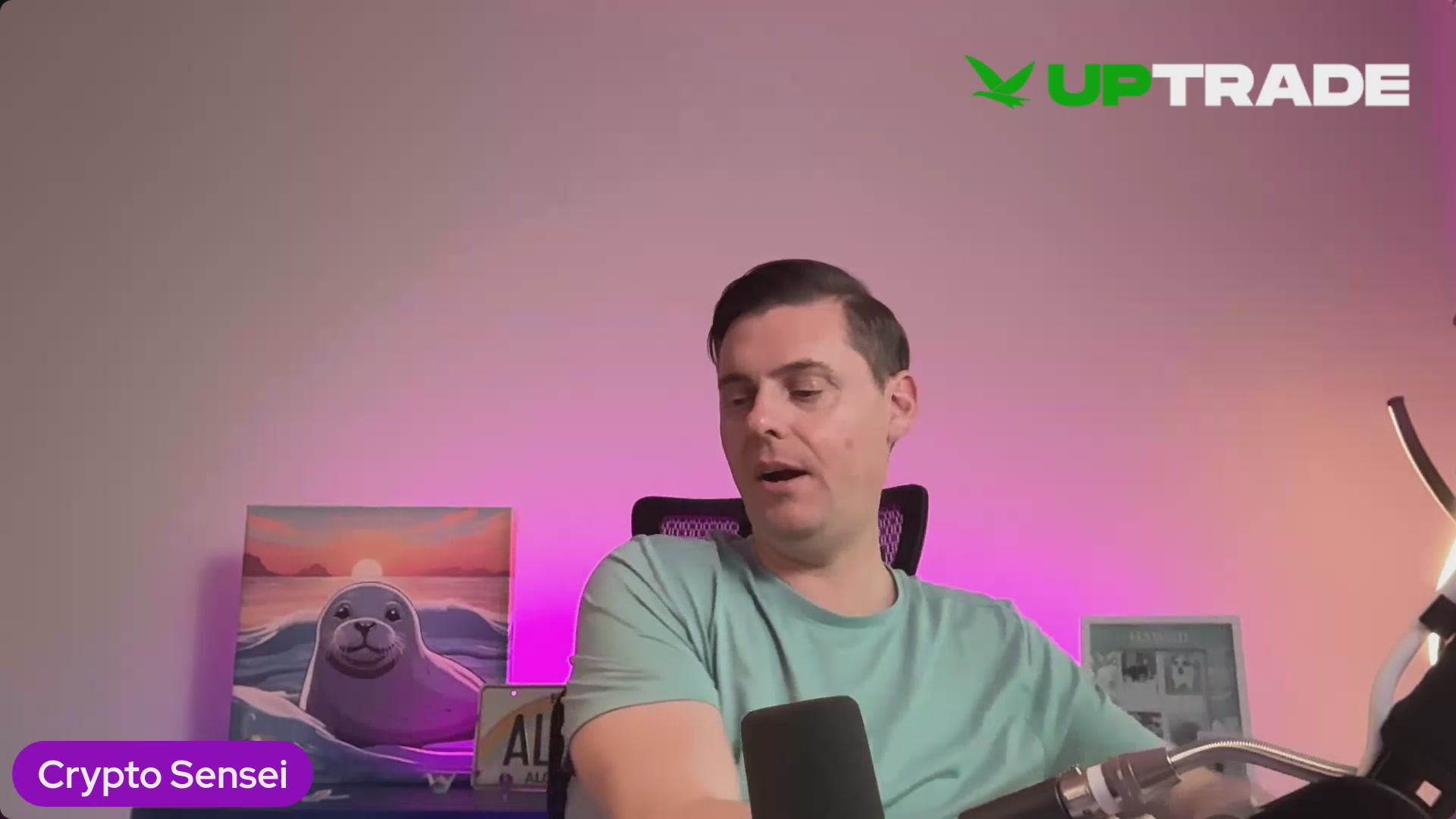 In this image, we see a man sitting down while talking into a microphone with the name 'UpTradeOfficial'. He appears to be discussing something related to cryptocurrency or tokenization, as he wears a green shirt that has the word "up" written on it. Behind him, there are several pieces of artwork displayed - specifically a painting featuring an octopus and another featuring a seal. The background is dominated by pink and purple lights, creating a unique atmosphere for this conversation.