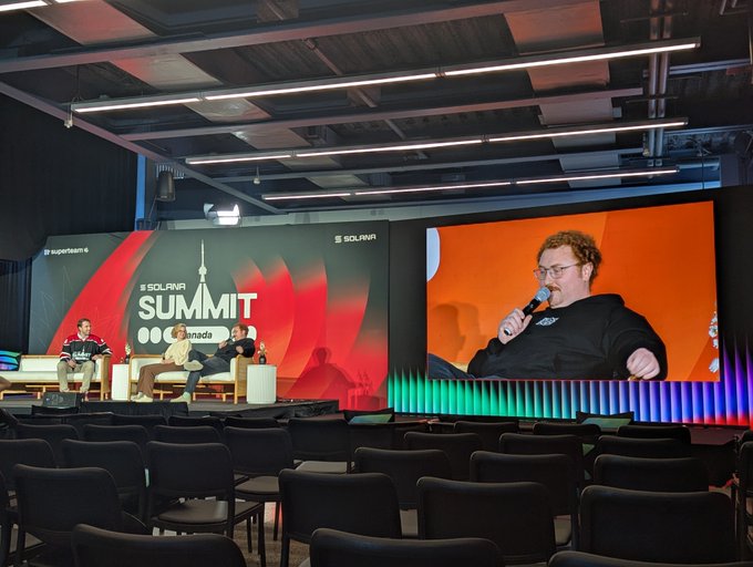 A panel discussion on stage at the SOLANA SUMMIT in Grenada, featuring three seated speakers on the left and a large screen displaying a close-up of a bearded man speaking into a microphone on the right, with empty chairs in the foreground.