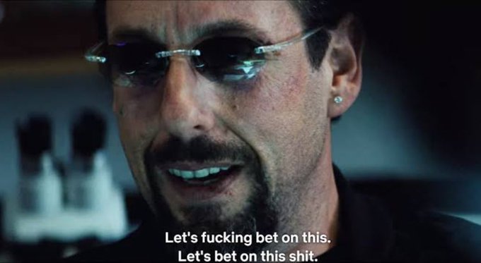 A close-up of a man with a goatee wearing sunglasses and an earring, smirking menacingly, with subtitles reading 'Let's fucking bet on this. Let's bet on this shit.'