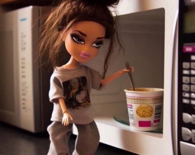 A Bratz doll with messy brown hair, wearing a printed t-shirt, posed as if cooking instant noodles with a spoon in front of an open microwave.