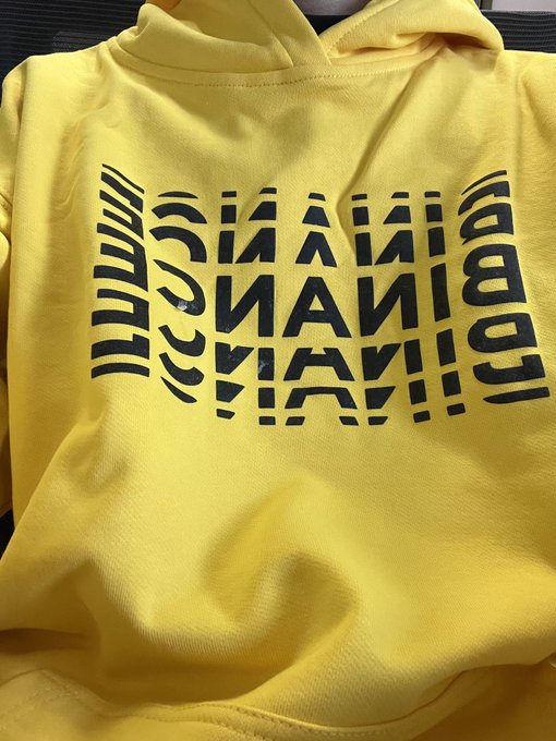 A yellow garment, likely a hoodie, featuring black screen-printed artwork with mirrored, abstract geometric typography.