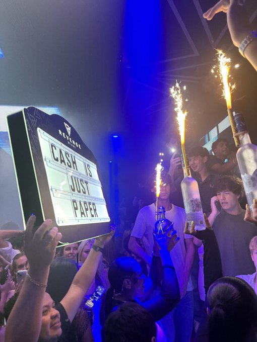 A lively nightclub scene where a person holds up a 'CASH IS JUST PAPER' sign at Shibuya LOVZ while others celebrate with sparkler-topped bottles under blue lighting.