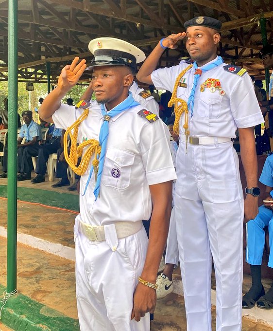 Two men in white naval-style uniforms with blue scarves and gold aiguillettes saluting, one wearing a white cap and the other a dark beret, standing in what appears to be an open-air pavilion with people seated in the background.