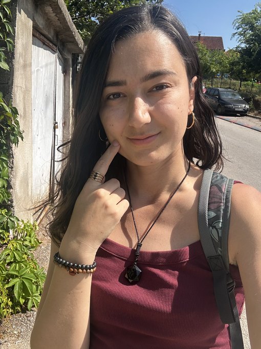A selfie of a young woman with dark hair wearing a maroon top, gold hoop earrings, rings, beaded bracelets, and a black pendant necklace, standing outdoors near a wall and gate with a car visible in the background.