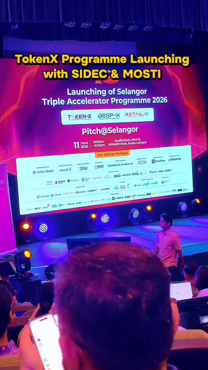 The image shows a large screen displaying information about an event, possibly a pitchx program launching or a similar gathering. The audience appears to be watching the presentation attentively from various positions around the room. There are multiple people present, including one person standing in front of the screen and others seated at tables. The setting seems to be a conference or exhibition where attendees can learn about new ideas and projects.