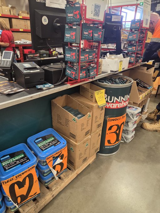 A photograph of a retail store checkout counter area displaying various hardware items, including blue buckets labeled 'ROGUE BATTERY', boxes of merchandise, a point-of-sale terminal, and a large 'BUNNINGS warehouse' cardboard barrel display.