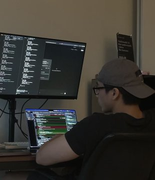 A person wearing a grey cap sits in a dark room facing two screens displaying trading charts and order book interfaces, with a handwritten note taped to the wall listing what appears to be trading rules or market levels.