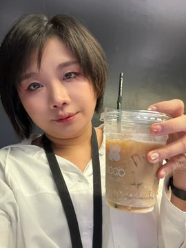 A short-haired woman in a white blouse smiles at the camera while holding up a plastic cup of iced coffee with the 'OGO' logo.