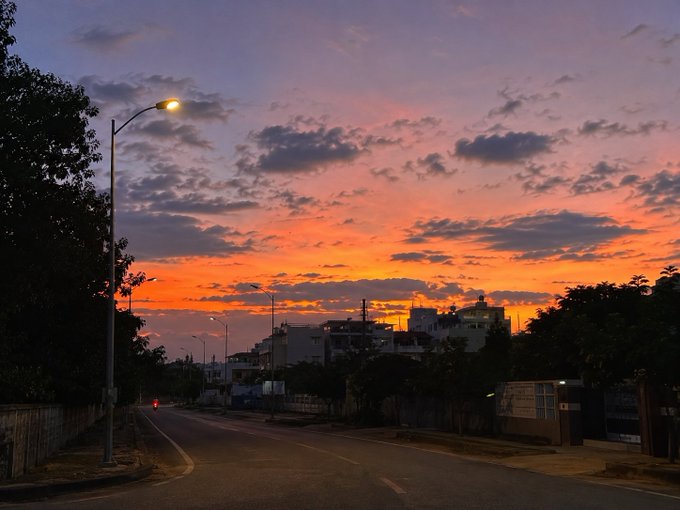 The image shows a street scene at sunset with a city skyline in the background. The sky has an orange hue as the sun sets behind the buildings. There are trees lining the street, adding greenery to the urban landscape. A street lamp is visible on one side of the road.