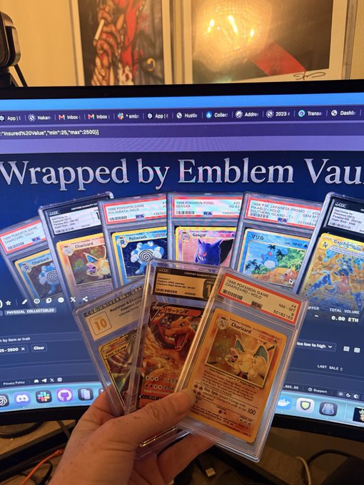 A hand holding several graded Pokémon cards, including Charizard (1st Edition), Magikarp, and Poliwhirl, in front of a monitor displaying a webpage titled 'Wrapped by Emblem Vault.'