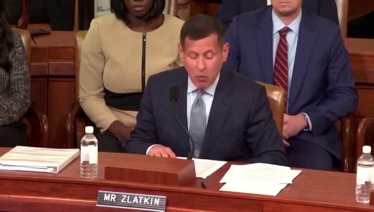 The image shows a man sitting at a wooden desk, wearing a suit and tie, while another person sits behind him. The scene appears to be set in a courtroom or a similar formal setting. There are two books placed on the table, one near the center of the table and the other towards the left side. A bottle is also present on the right side of the desk.

The image includes text that reads "NO TAXES ON BITCOIN PAYMENTS Coinbase just urged Congress to remove taxes on Bitcoin payments." This message seems to be related to the man sitting at the desk, possibly discussing or advocating for the removal of tax implications associated with Bitcoin payments.