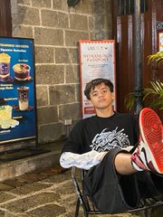 A young person sits on a chair in front of a stone wall, casually lifting one foot to show the red sole of their shoe.