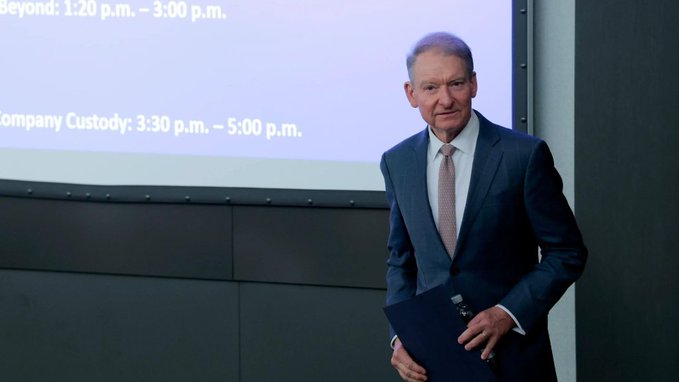 An older man in a dark suit and tie stands holding a folder in front of a presentation screen displaying text including 'Beyond: 1:20 p.m. – 3:00 p.m.' and 'Company Custody: 3:30 p.m. – 5:00 p.m.'