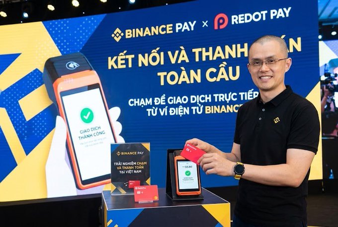 A smiling man in a black polo shirt demonstrates a Binance Pay x Redot Pay payment terminal against a blue and yellow backdrop with Vietnamese text 'KẾT NỐI VÀ THANH TOÁN TOÀN CẦU', with a sign reading 'Binance Pay - Triển khai thành công tại Việt Nam'.