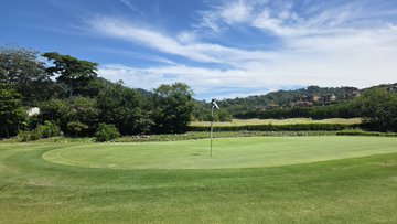 A golf green with a pin nestled against rolling, tree-lined hills under a bright blue sky with wispy clouds, capturing the scenic "links" course referenced in the post. The image evokes the calm, picturesque setting of the golfer's successful round featuring 2 birdies and a back-nine par.