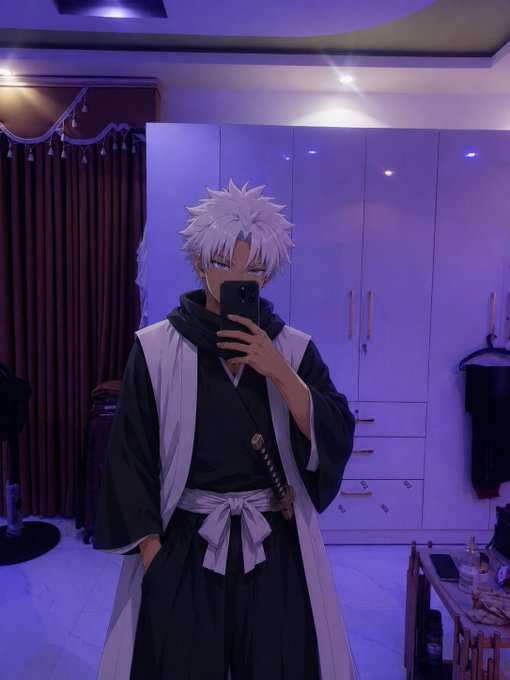 A person cosplaying as an anime character (resembling Toshiro Hitsugaya from Bleach) with spiky white hair, a dark kimono with white haori, and a sword, taking a mirror selfie in a purple-lit room.