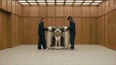 An art gallery scene showing two men in suits examining a classical stone sculpture of a seated figure displayed on a pedestal in a minimalist wood-paneled room.