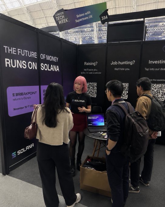 The image shows a group of people gathered around a booth with a laptop, possibly discussing or learning about Solana, a cryptocurrency platform. There are four individuals standing near the booth, including two women and two men. One woman appears to be giving a presentation on the platform while the others listen attentively. The backdrop features a banner that reads "The Future of Money Runs On Solana" and "Superstars with UK jobs hunting? Investing? Job hunting?" This suggests that the event is related to the cryptocurrency industry or financial technology, possibly at a Tech Week gathering.

There are also two handbags visible in the scene, one near each person standing around the booth. The overall atmosphere seems to be professional and focused on learning about Solana and its potential applications for various industries.
