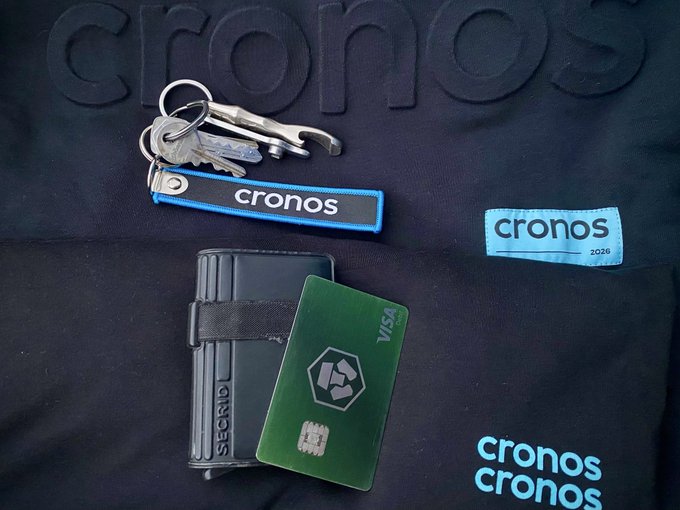A flat-lay photo of Cronos-branded merchandise on black fabric, including keys on a 'cronos' lanyard, a Secrid cardholder with a green Visa card, and 'cronos 2026' labels.