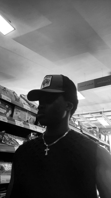 A black and white photo of a man wearing a cap, sunglasses, and a cross necklace standing in what appears to be a grocery store aisle.