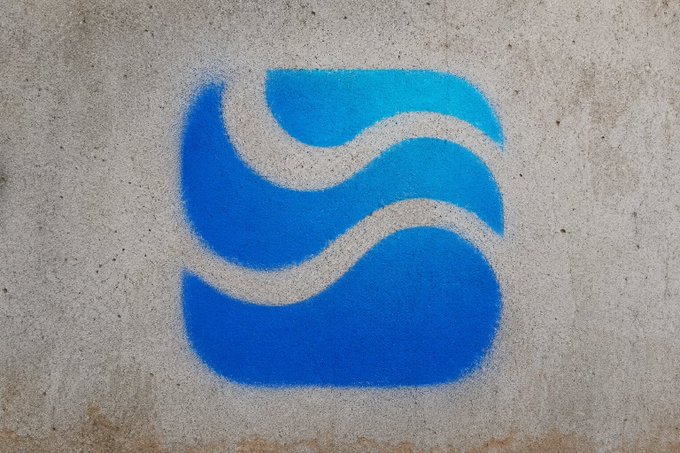 A stylized blue wave or 'S' logo is spray-painted in gradient blue tones on a textured gray concrete wall.