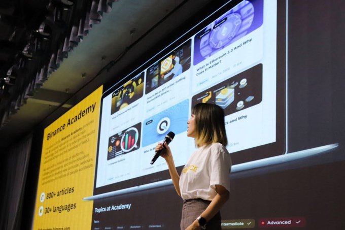 A woman in a white t-shirt speaks into a microphone on stage, presenting in front of a large screen displaying a Binance Academy interface with '800+ articles' and '30+ languages' visible.