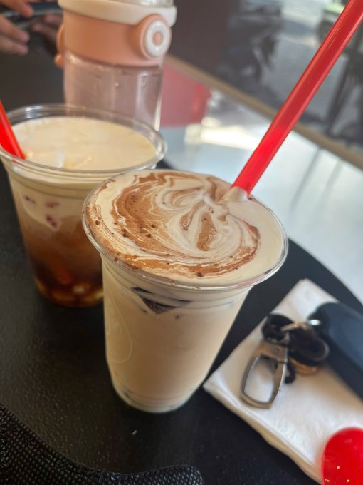 A close-up photograph shows two iced coffee drinks on a dark table, one a latte with a swirled cinnamon foam art top and a red straw, the other a darker coffee with red boba or jelly bits, alongside a pair of sunglasses on a napkin.