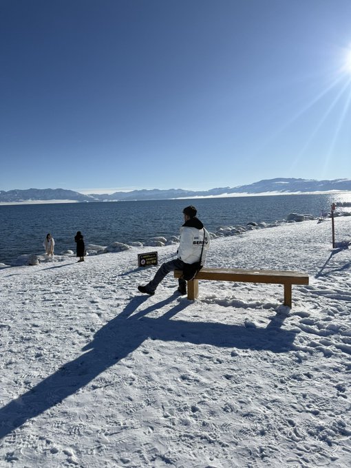A person sits alone on a wooden bench on a snowy shoreline beside a large body of water, with mountains visible in the distance under a bright blue sky.
