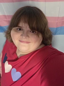 A young person with brown hair poses in front of a pink, blue, and white striped flag, wearing a red shirt decorated with pink, white, and purple hearts. The dramatic caption "What the FUCK is THAT???!??!?" seems to humorously react to the heart-patterned shirt or the bold trans flag backdrop.