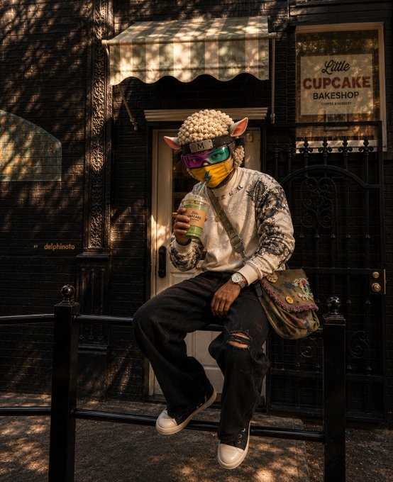 A person in an elaborate streetwear outfit featuring a sheep mask, colorful ski goggles, and holding a coffee cup, sitting on a railing in front of 'Little CUPCAKE BAKESHOP'.